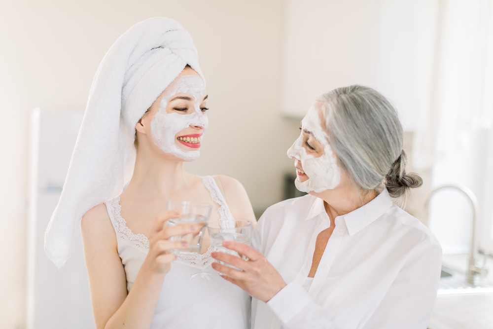 Healthy lifestyle and family spa. Horizontal shot of two Caucasian pretty women, grandmother and granddaughter, having beauty procedures with facial clay mask at home and holding glasses of water Healthy lifestyle and family spa. Horizontal shot of two Caucasian pretty women, grandmother and granddaughter, having beauty procedures with facial clay mask at home and holding glasses of water.