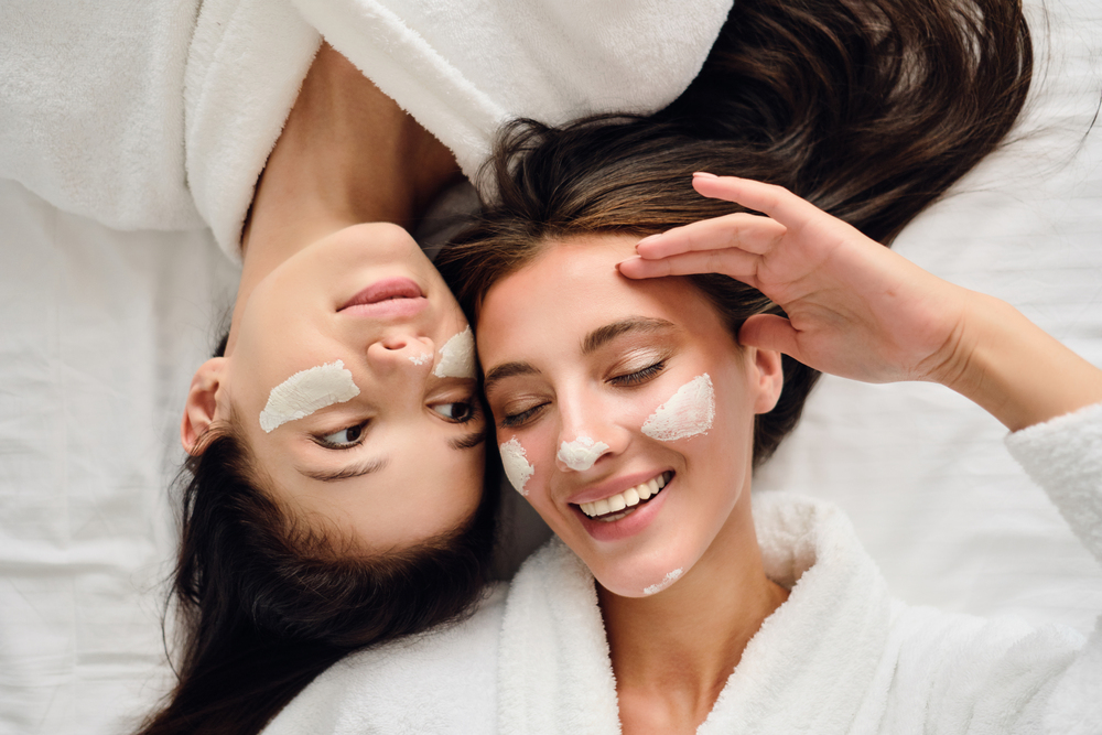 Two young pretty smiling women with dark hair in white bathrobes happily lying near in bed with cosmetic mask on faces in modern cozy hotel